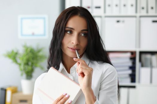 Portrait of woman holding silver pen on mouth, thinking about new ideas to journal. Female in suit at workspace with organizer diary. Creativity concept