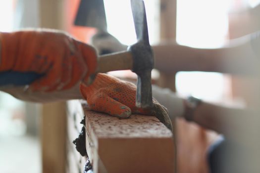 Close-up of handyman hammering on red brick with tool, worker wear gloves at work. Busy on new building object, repair and fix. Renovation, build concept