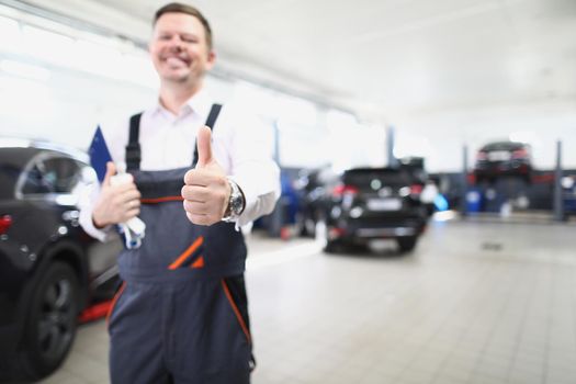 Portrait of happy worker in uniform show thumbs up, well done service job. Fixed car after maintenance in garage. Pit stop, service station, fixing concept