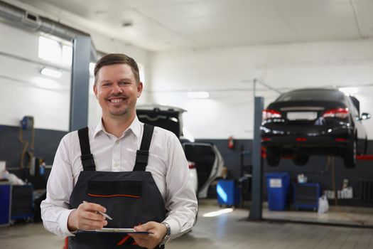 Portrait of smiling qualified maintenance center worker in uniform, man posing on cars background at work. Male propose high service. Pit stop, fix concept