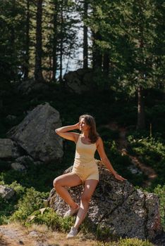 Young woman tourist sitting on the mountain peak and beautiful mountains. Landscape with sporty girl, green forest, hills , sky, lake. Travel and tourism. Hipster girl in the mountains. Stylish woman in checkered shirt sitting on stones in forest. Wanderlust concept. Hiking and travelling in summer. mysterious wood. Beautiful nature