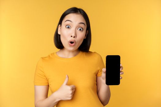 Excited asian woman showing mobile phone screen, thumbs up, like smth good, recommending smartphone application, standing over yellow background.