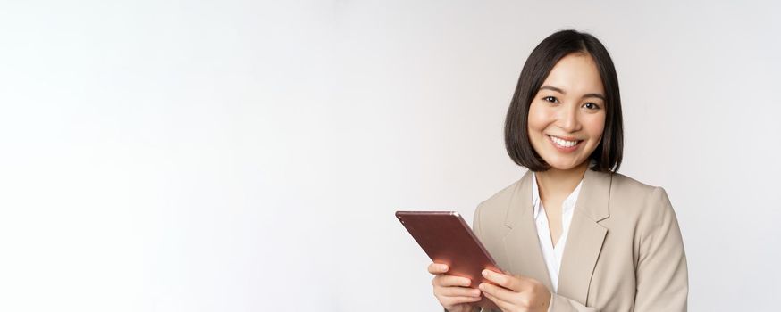 Image of asian businesswoman, saleswoman holding digital tablet and smiling, working with gadget, standing in suit over white background.