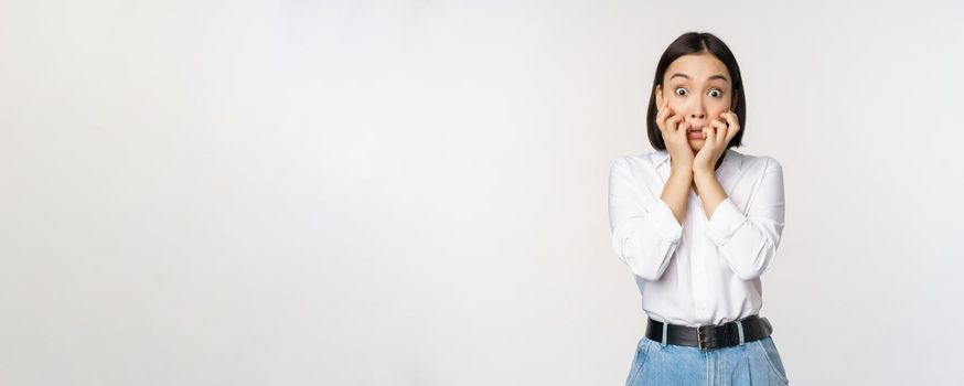 Portrait of scared asian woman watching smth scary, biting fingers on hands and looking at camera frightened, standing over white background.