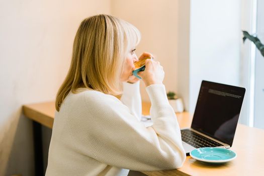 Mature adult woman sitting in cafe, drinking coffee mug and working online on laptop. Businesswoman in eyeglasses typing on notebook computer, in coworking space in roasters coffee shop.