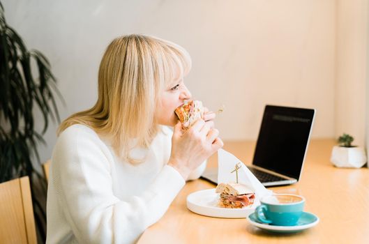 Mature adult woman sitting in cafe with coffee mug eating sandwiches and working online at laptop. Businesswoman in typing on notebook computer, in coworking space in roasters coffee shop.