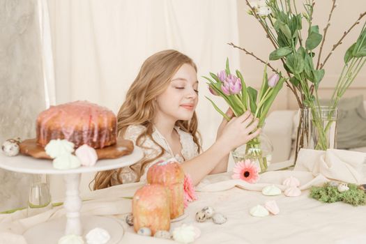A girl with long hair in a light dress is sitting at the Easter table with cakes, spring flowers and quail eggs. Happy Easter celebration