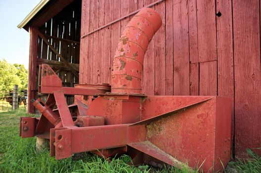 Giant Vintage Antique Rusty Red Snowblower attachment for Tractor beside a Red Barn in the Summer. High quality photo