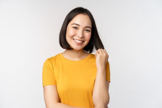 Beautiful romantic asian girl, smiling and playing with hair, looking happy at camera, standing in yellow t-shirt over white background.