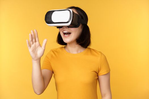 Girl in VR. Beautiful young asian woman, using virtual reality glasses and playing, chatting virtually, standing over yellow background.