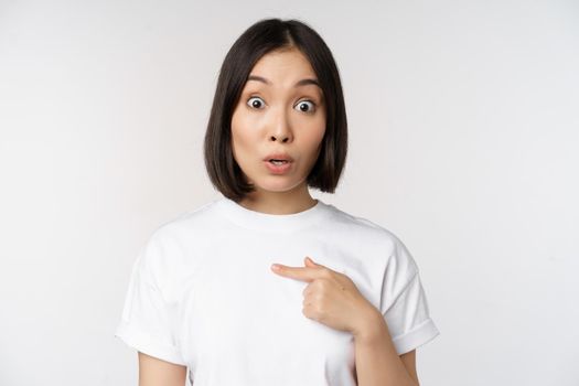 Portrait of surprised asian girl student, pointing finger left, raising eyebrows amazed, impressed by big deal, offer ahead, standing over white background.