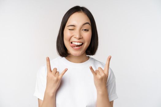 Sassy asian girl shouting, enjoying concert or festival, showing rock on, heavy metal sign, having fun, standing over white background.