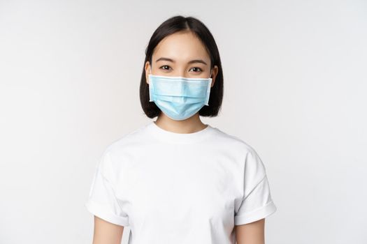 Health and covid pandemic concept. Smiling asian girl wearing medical face mask, looking happy and confident, protecting herself from coronavirus, white background.