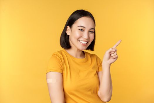 Vaccination and covid-19 pandemic concept. Smiling korean woman with band aid on shoulder after coronavirus vaccine shot, pointing at banner with vaccinating campaign.