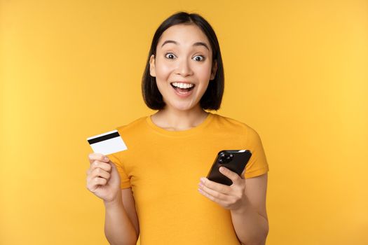 Online shopping. Cheerful asian girl holding credit card and smartphone, paying, order with mobile phone, standing over yellow background.