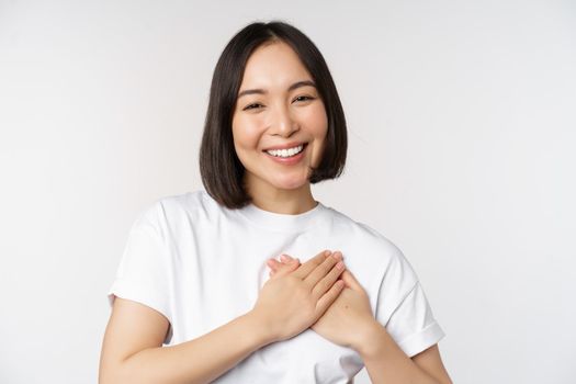 Close up of smiling korean woman holding hands on heart, care and love concept, feel affection, tenderness or heartwarming feeling, standing over white background.