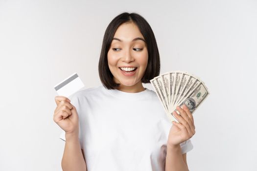 Portrait of asian woman smiling, holding credit card and money cash, dollars, standing in tshirt over white background.