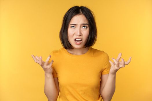 Image of angry asian woman, shouting and cursing, looking outraged, furious face expression, standing over yellow background.