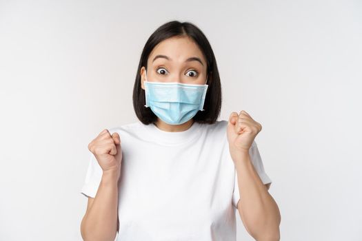 Covid-19, healthcare and medical concept. Enthusiastic asian woman in medical face mask, dancing and celebrating, winning, achieve goal, standing over white background.