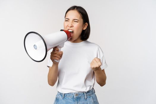 Confident asian woman shouting in megaphone, screaming and protesting. Girl activist using speaker to speak louder, standing over white background.