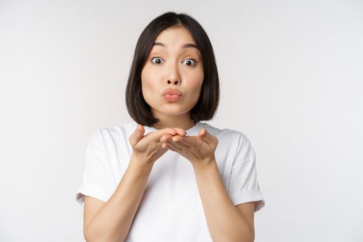 Lovely young asian woman, sending air kiss and looking coquettish at camera, standing in tshirt over white background.