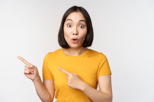 Portrait of smiling asian brunette girl in yellow tshirt, pointing fingers left, showing copy space, promo deal, demonstrating banner, standing over white background.