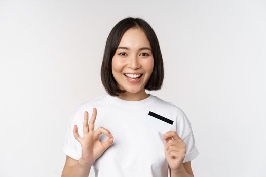 Smiling korean girl showing okay sign and recommending credit card of copy space bank, standing in tshirt over white background.