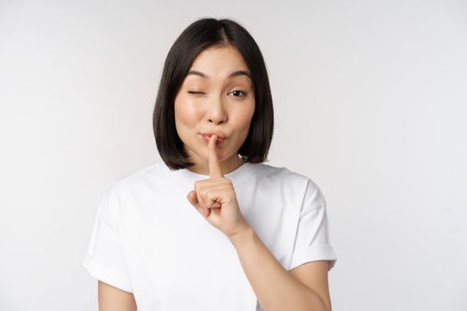 Close up portrait of young beautiful asian girl shushing, has secret, keep quiet silence gesture, press finger to lips, standing in tshirt over white background.