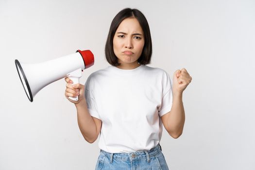 Angry asian girl activist, holding megaphone and looking furious, protesting, standing over white background. Copy space