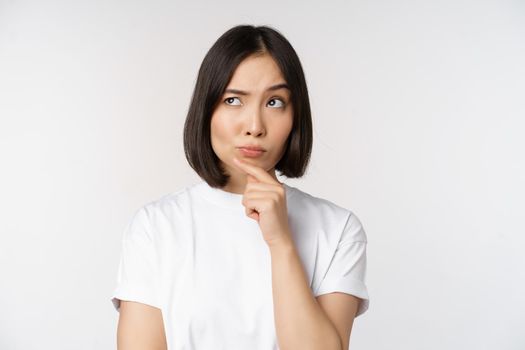 Thoughtful asian woman looking aside, pondering, making assumption, thinking or choosing smth, standing over white background.