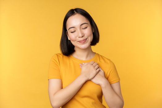 Romantic korean girl, asian woman holding hands on heart, smiling with care and tenderness, standing over yellow background.