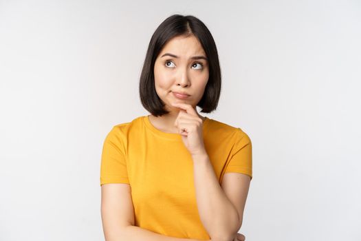 Image of thinking asian woman, looking aside and pondering, making decision, standing in yellow tshirt over white background.