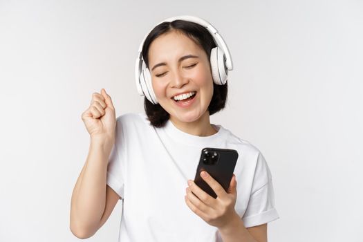 Beautiful modern asian girl, listening music in headphones, holding mobile phone, using smartphone app, standing over white background.