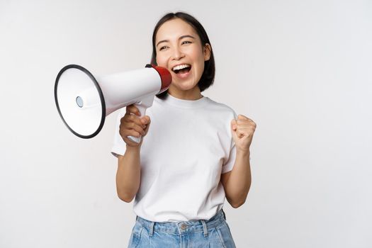 Asian girl shouting at megaphone, young activist protesting, using loud speakerphone, making announcement, white background. Copy space