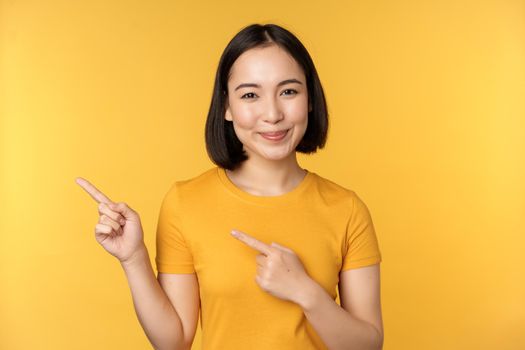 Smiling asian woman pointing fingers left, showing advertisement on empty copy space, standing over yellow background.