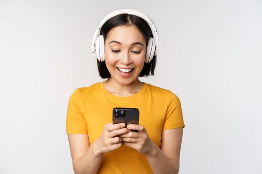 Cute japanese girl in headphones, looking at mobile phone and smiling, using music app on smartphone, standing against white background.