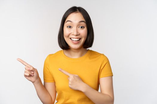 Portrait of smiling asian brunette girl in yellow tshirt, pointing fingers left, showing copy space, promo deal, demonstrating banner, standing over white background.