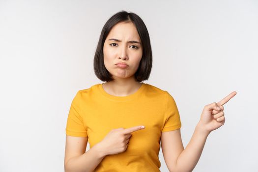 Portrait of disappointed, moody asian girl, pointing fingers right and looking at smth unfair, complaining, standing over white background.