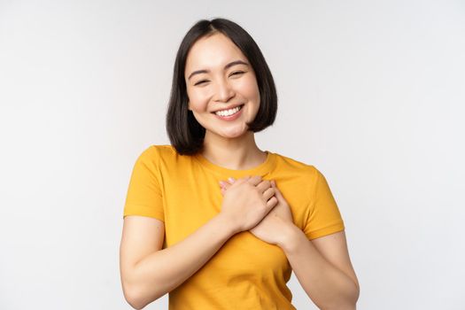 Romantic asian girfriend, holding hands on heart, smiling with care and tenderness, standing in yellow tshirt over white background.