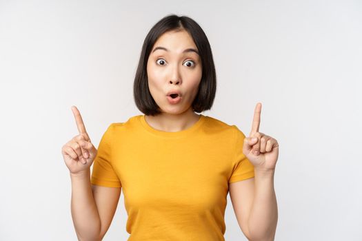 Portrait of impressed asian girl, pointing fingers up, say wow, looking amazed at camera, standing over white background. Copy space