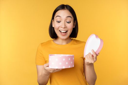 Portrait of excited asian woman, open gift box with surprised happy face, standing over yellow background.