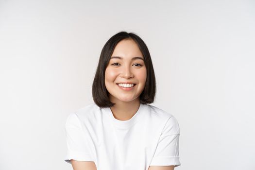 Beautiful korean girl smiling, white teeth, looking lovely at camera, standing in white tshirt over studio background.