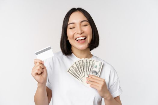 Portrait of asian woman smiling, holding credit card and money cash, dollars, standing in tshirt over white background.