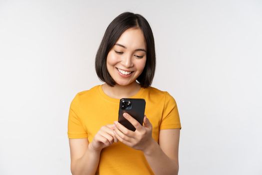 People and technology concept. Smiling asian girl using smartphone, texting on mobile phone, standing against white background.