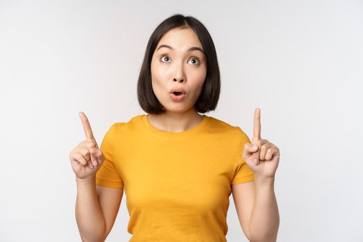 Happy beautiful asian girl showing advertisement, pointing fingers up, standing over white background.