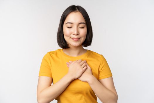 Romantic asian girfriend, holding hands on heart, smiling with care and tenderness, standing in yellow tshirt over white background.