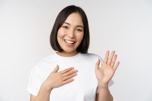 Portrait of beautiful korean girl raising hand, introduce herself, put hand on heart, greeting, standing over white background.