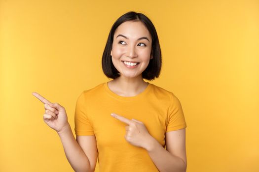 Portrait of happy asian girl pointing fingers and looking left, smiling amazed, checking out promo banner, showing advertisement against yellow background.