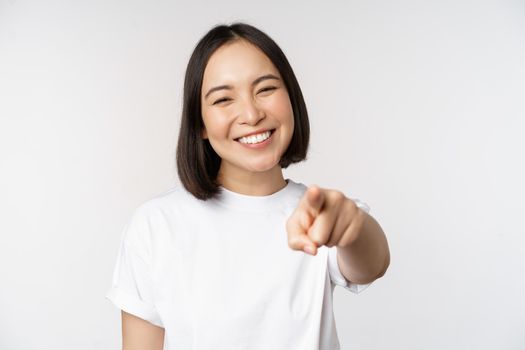 Portrait of happy smiling asian woman with white teeth, pointing finger at camera, choosing you, congratulating, standing over white background.