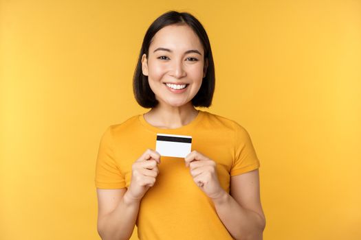 Beautiful asian woman showing credit card and smiling, recommending bank service, standing over yellow background. Copy space